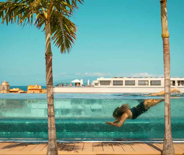 En person svømmer under vandet i en gennemsigtig pool, med bygninger og en palme mod en blå himmel i baggrunden. (AI-genereret tekst)