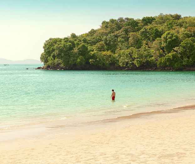 En person i røde badebukser står i vandet ved en strand nær frodig skov. (AI-genereret tekst)