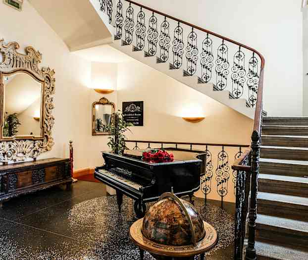 En trappe med smedejernsgelænder, en globus, et sort flygel med røde blomster på samt et dekorativt spejl. (AI-genereret tekst)