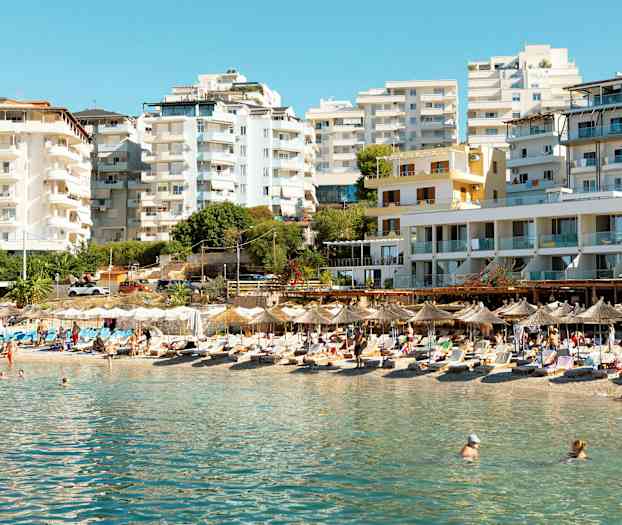 En strand med parasoller og liggestole foran højhuse. (AI-genereret tekst)