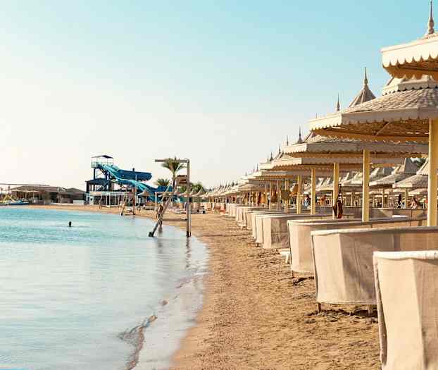 En sandstrand med liggestole og parasoller samt vandrutsjebaner i baggrunden. (AI-genereret tekst)