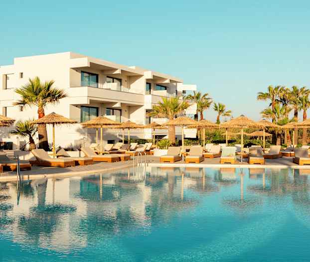 Hotel med hvid facade bag en swimmingpool og liggestole med parasoller. (AI-genereret tekst)