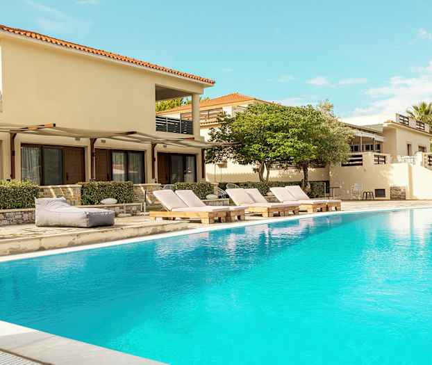 Udendørs swimmingpool foran bygninger med liggestole og omkringliggende vegetation. (AI-genereret tekst)