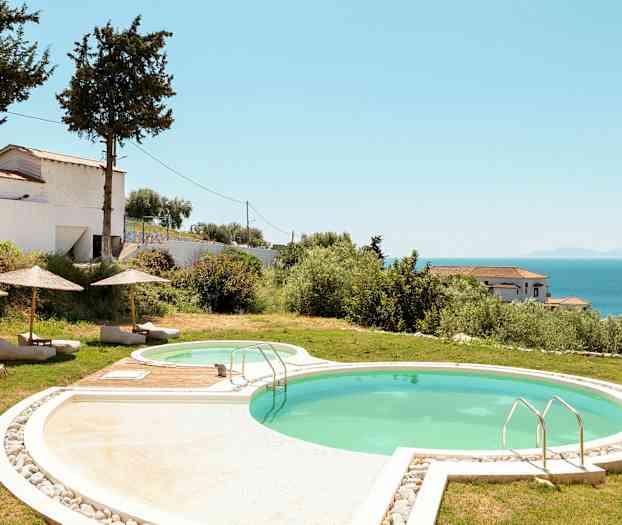 En udendørs pool med liggestole og parasoller, omgivet af græs og vegetation. Havet og bygninger ses i baggrunden. (AI-genereret tekst)