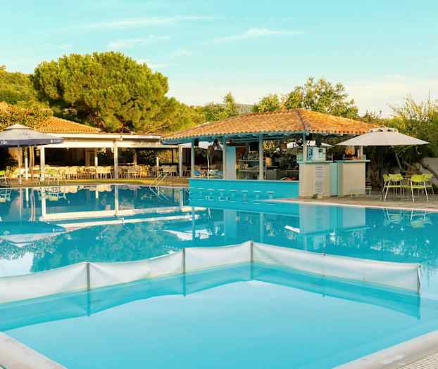 En udendørs pool med bardiske i vandet, omgivet af borde og stole samt grønne områder. (AI-genereret tekst)