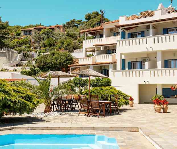 Hvid bygning med altaner, frodig have og pool i forgrunden. (AI-genereret tekst)