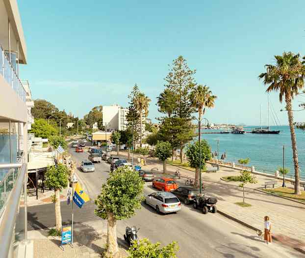 En bygade, der går ved siden af havet, med biler, palmer og nogle bygninger i baggrunden. (AI-genereret tekst)