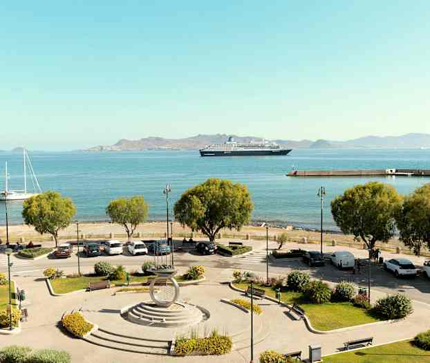 Udsigt over en havn med parkeringsplads og træer i forgrunden, både og et krydstogtskib på det blå hav, samt bjerge i baggrunden. (AI-genereret tekst)