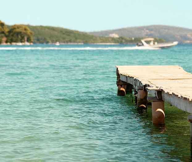 En træbro, der strækker sig ud i klart vand, med en motorbåd og frodige bakker i baggrunden. (AI-genereret tekst)
