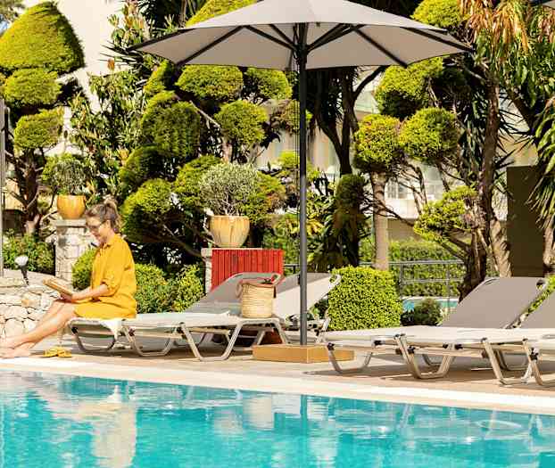 En kvinde i en gul kjole sidder med en åben bog ved en pool, omgivet af liggestole og parasoller. (AI-genereret tekst)