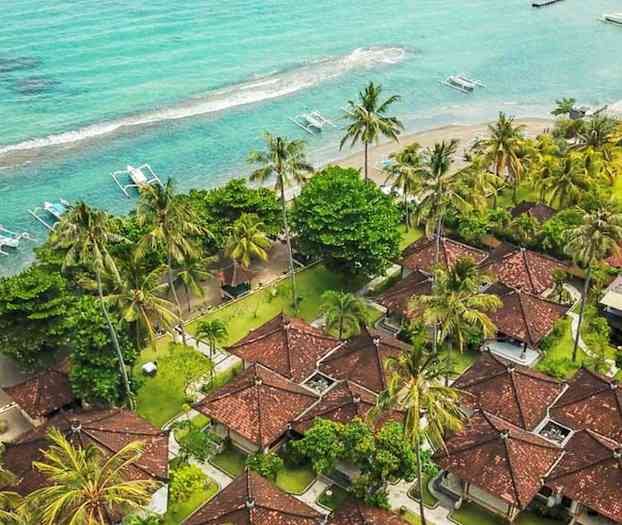 Luftfoto af et strandresort med røde hustage, omgivet af palmer og tæt på et turkisblåt hav. (AI-genereret tekst)