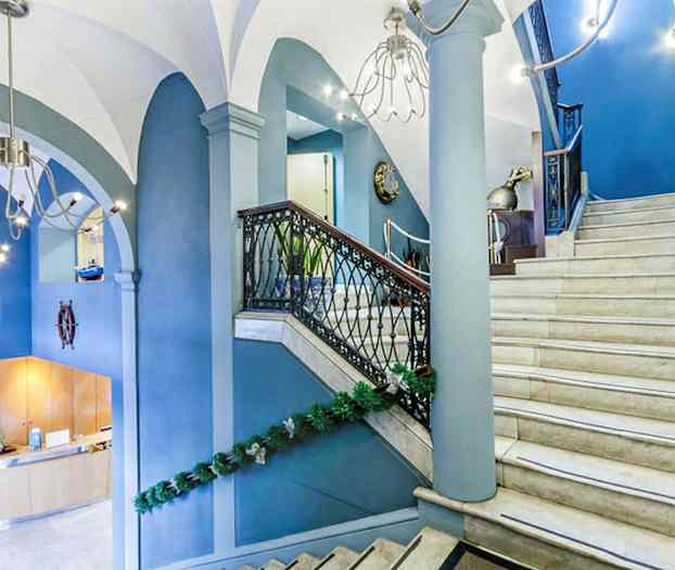Et interiørbillede af en hall med en trappe i lys marmor og blå vægge, dekoreret med grønne guirlander. Trappen har gelænder i sort metal og trædetaljer. (AI-genereret tekst)