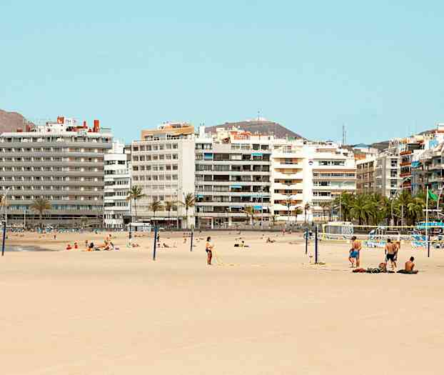 Strand med sand og mennesker, der soler sig og spiller volleyball foran flere højhuse. (AI-genereret tekst)