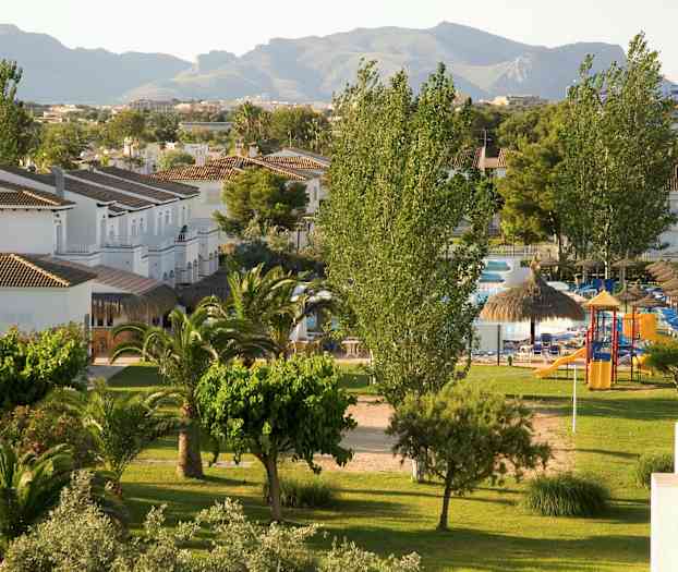 Udsigt over et grønt område med træer, græsplæne og små hvide bygninger samt baggrund med bjerge. (AI-genereret tekst)