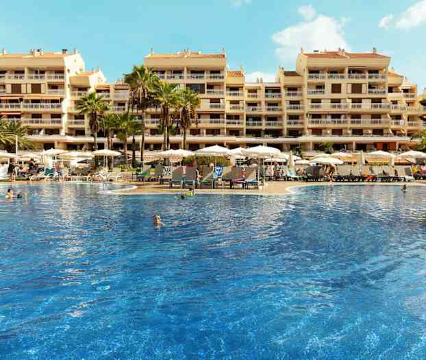 En stor pool omgivet af liggestole og parasoller foran en hotelbygning med flere etager. (AI-genereret tekst)