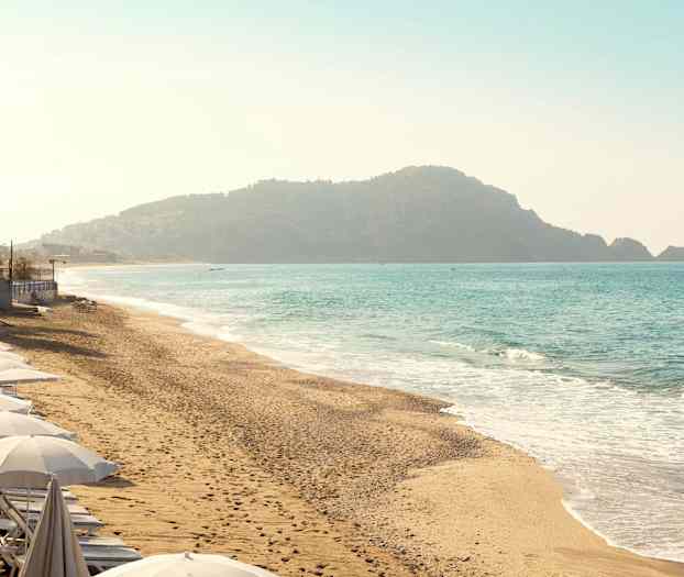 Strand med parasoller langs kystlinjen og baggrund med bjerge ved havet. (AI-genereret tekst)