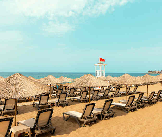 Strand med liggestole og parasoller samt et livreddertårn med rødt flag. (AI-genereret tekst)