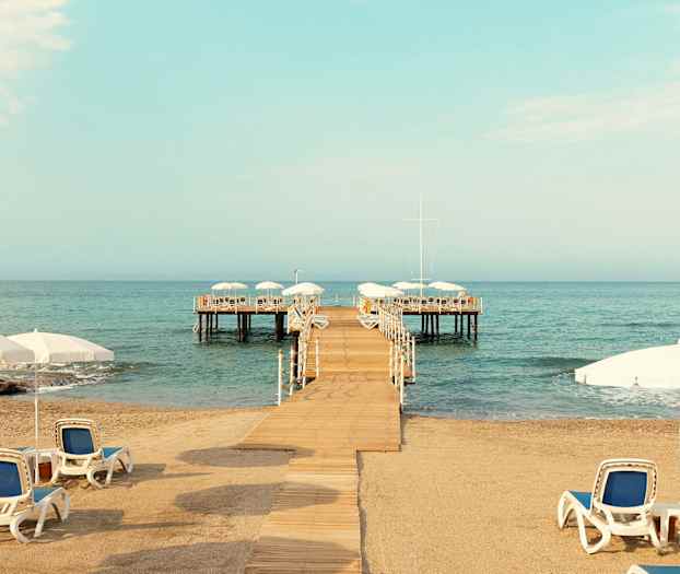 Sandstrand med liggestole og parasoller, en bro der fører ud til en platform over vandet. (AI-genereret tekst)