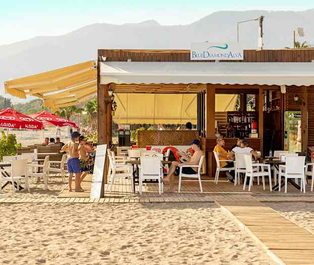 Strandrestaurant med udendørs servering og liggestole, bjerge i baggrunden. (AI-genereret tekst)