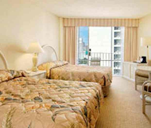 Et hotelværelse med to senge med blomstret sengetæppe, et lille møbel med en kaffemaskine og en balkon med udsigt over byen. (AI-genereret tekst)