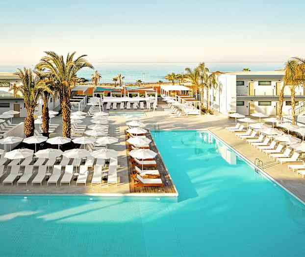 En hotelpool omgivet af solsenge og parasoller, med palmer og havudsigt i baggrunden. (AI-genereret tekst)