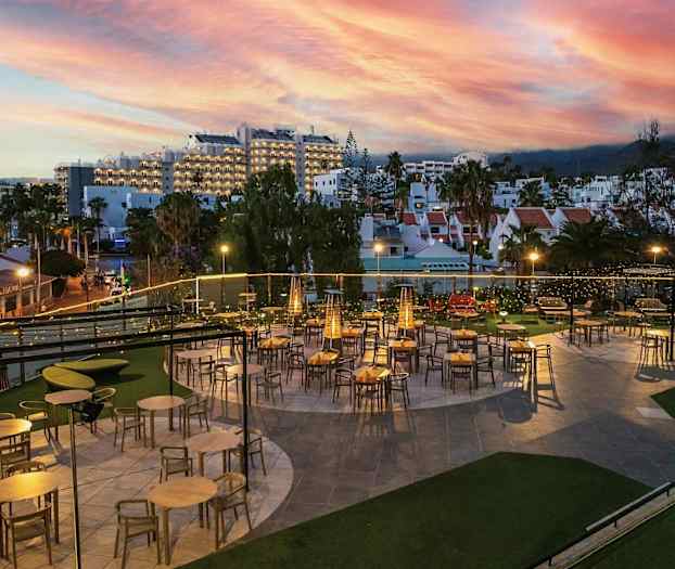 En udendørsrestaurant med borde og stole under aftenbelysning. I baggrunden ses bygninger og en farverig solnedgang. (AI-genereret tekst)
