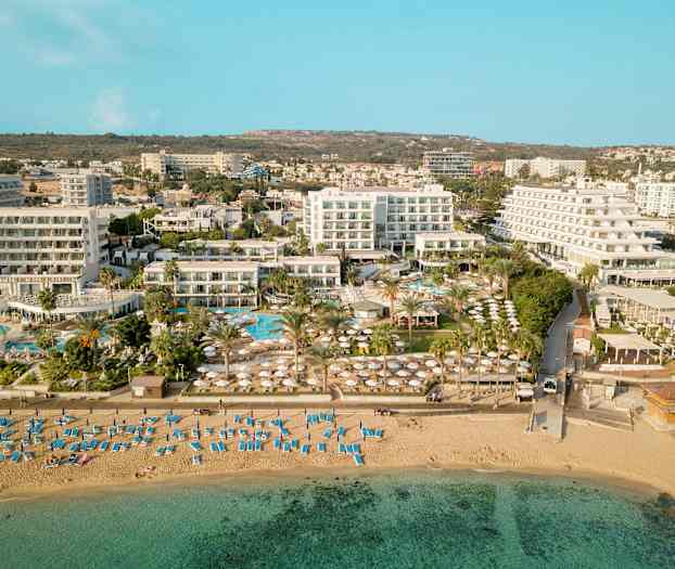 Luftfoto, der viser en strand med liggestole og parasoller samt hotelbygninger og poolområde i baggrunden. (AI-genereret tekst)
