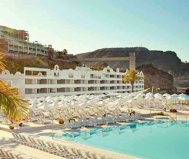 Hotelbygning med hvide facader, liggestole og parasoller ved en stor pool, samt klippeformationer i baggrunden. (AI-genereret tekst)