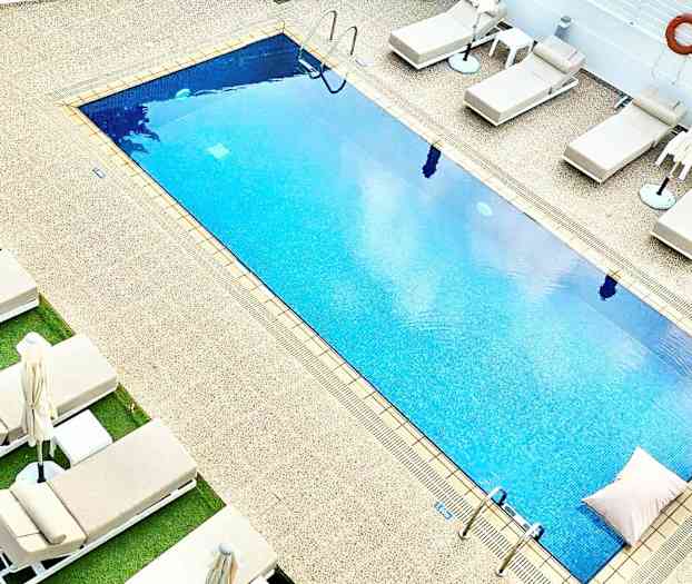 Rektangulær pool med blåt vand omgivet af liggestole og parasoller. (AI-genereret tekst)
