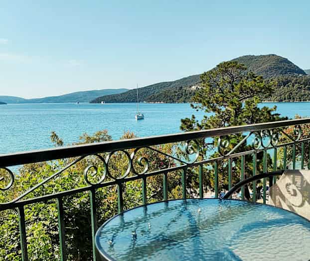 Et glasbord og stole på en altan med udsigt over havet, grøn beplantning og bjerge i baggrunden. (AI-genereret tekst)