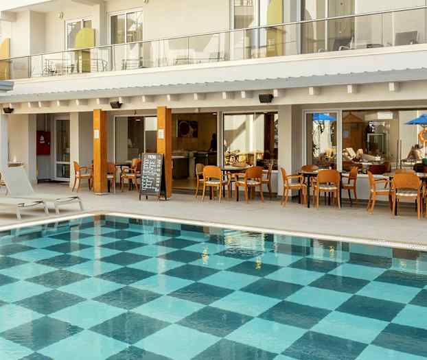 Udendørs pool med ternet bundmønster foran hotelbygning med altaner og restaurant. (AI-genereret tekst)