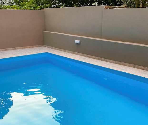 Udendørs pool med klart blåt vand, omgivet af en beige mur og grønt i baggrunden. (AI-genereret tekst)