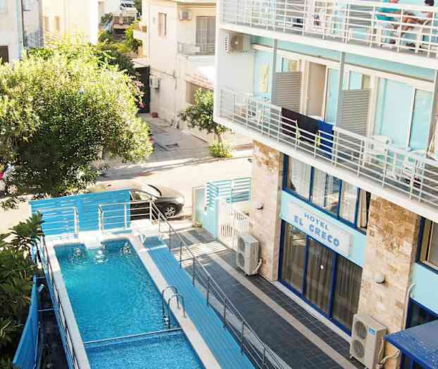 En swimmingpool med blåt vand omgivet af hegn, beliggende ved et hotel med blå facade. (AI-genereret tekst)