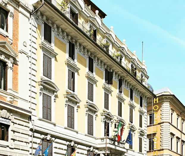 Facade af et hotel med flere etager, dekoreret med flag og omgivet af biler og motorcykler. (AI-genereret tekst)