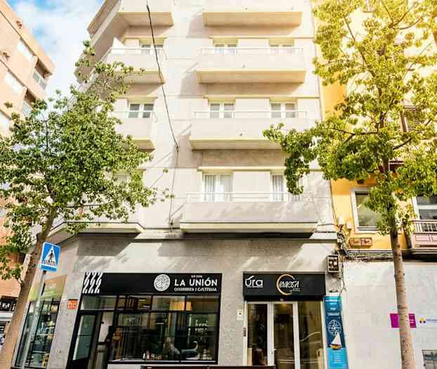 Boligbygning med altaner, butiksfacader på gadeplan og lidt grønne områder omkring. (AI-genereret tekst)