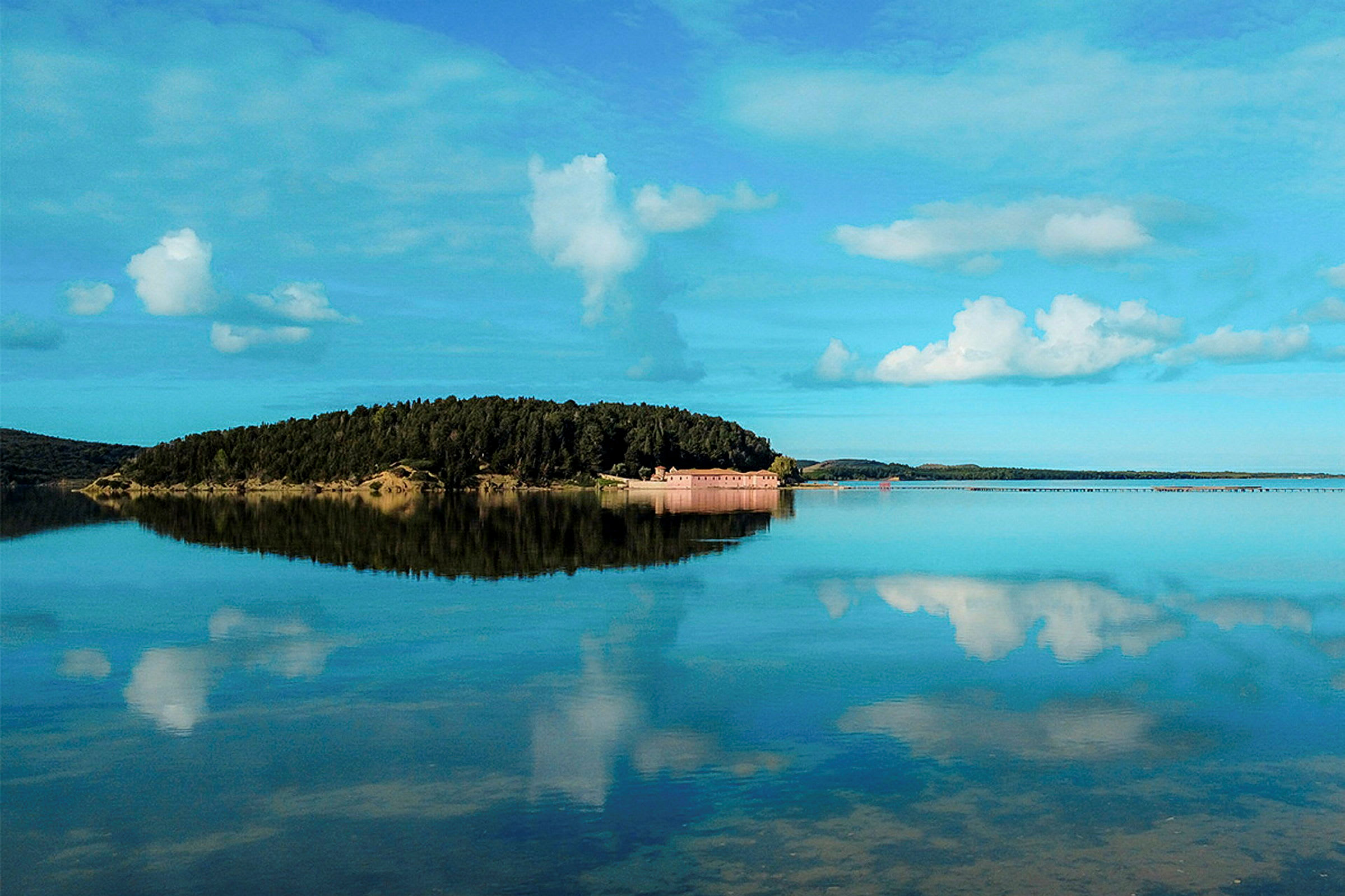 En ø med skov og bygninger omgivet af stille vand, med en klar himmel og skyer. (AI-genereret tekst)