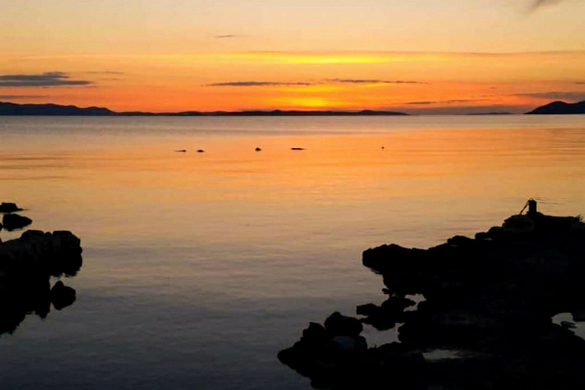 Solnedgang over et stille hav med silhuetter af klipper i forgrunden. (AI-genereret tekst)