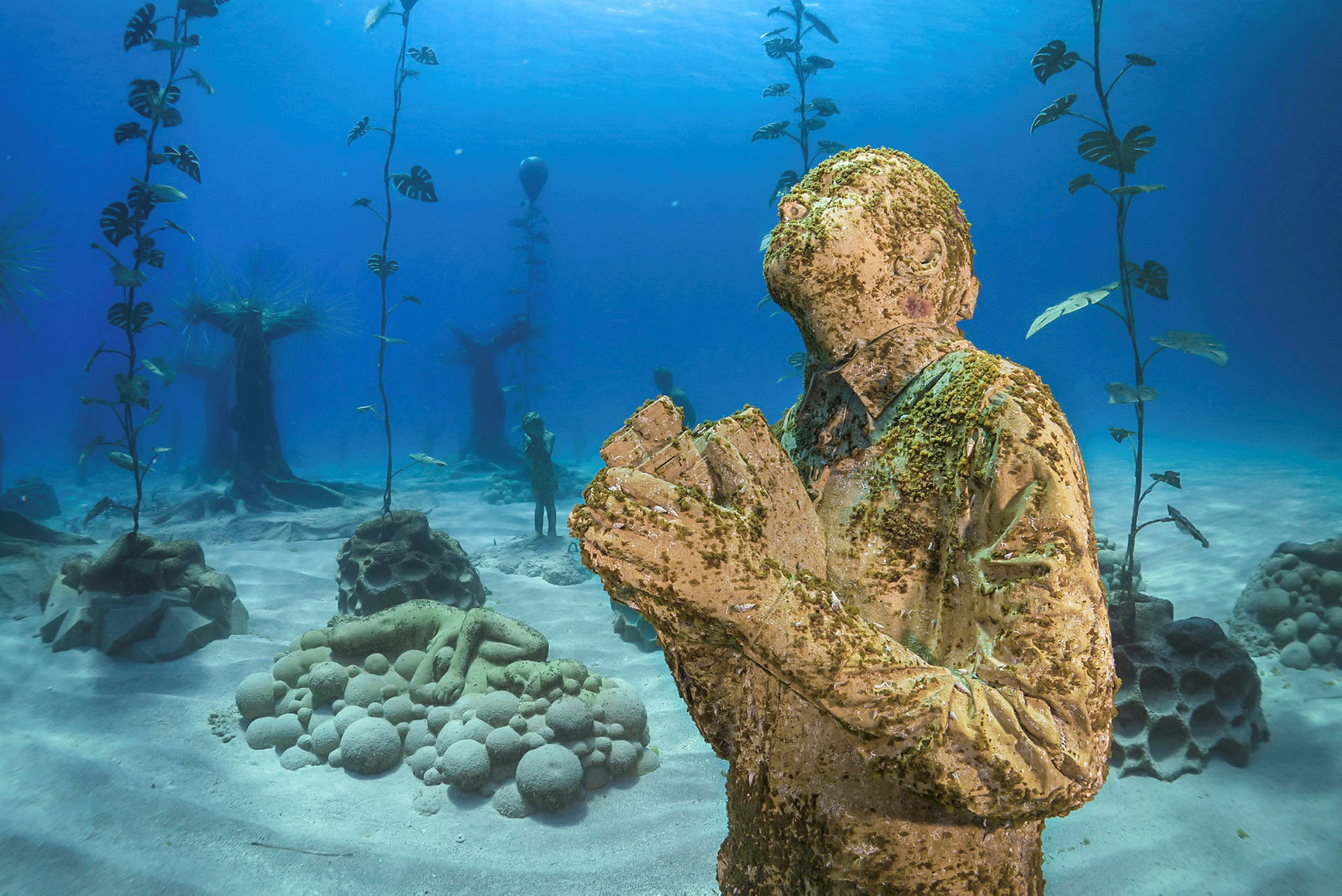 Undervandsbillede, der viser en statue af en person i en bedeposition, omgivet af undervandsplanter og andre marine skulpturer. (AI-genereret tekst)