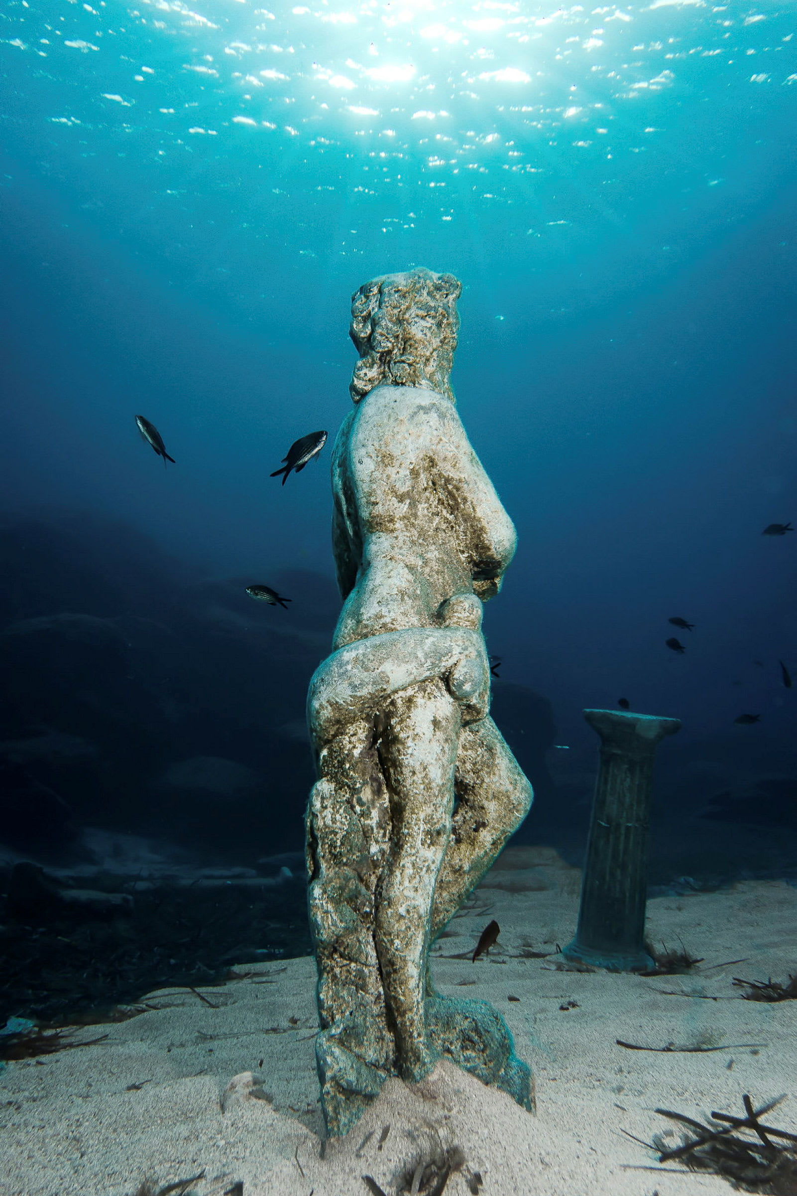 Et undervandsbillede af en gammel statue omgivet af sand og små fisk. (AI-genereret tekst)