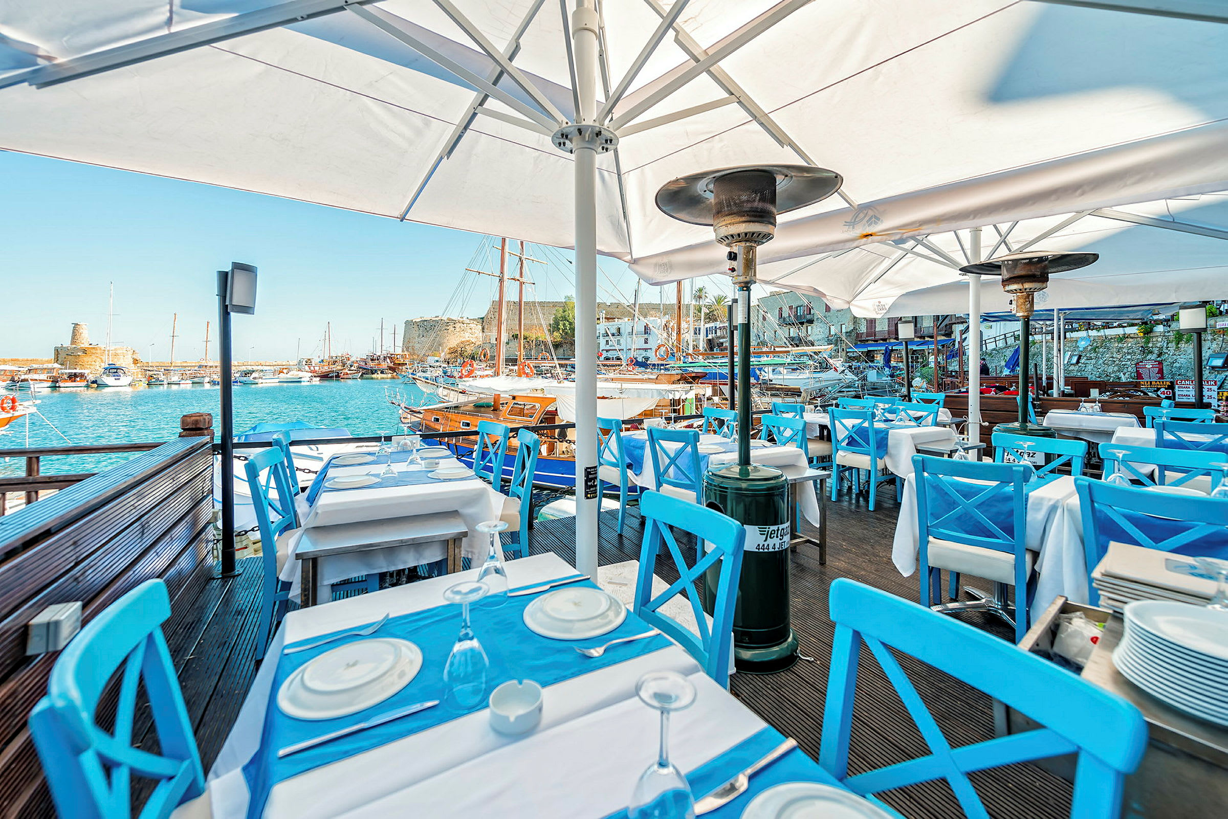 En restaurant med blå stole og hvide borde ved en havn. Sejlskibe er synlige i baggrunden. (AI-genereret tekst)