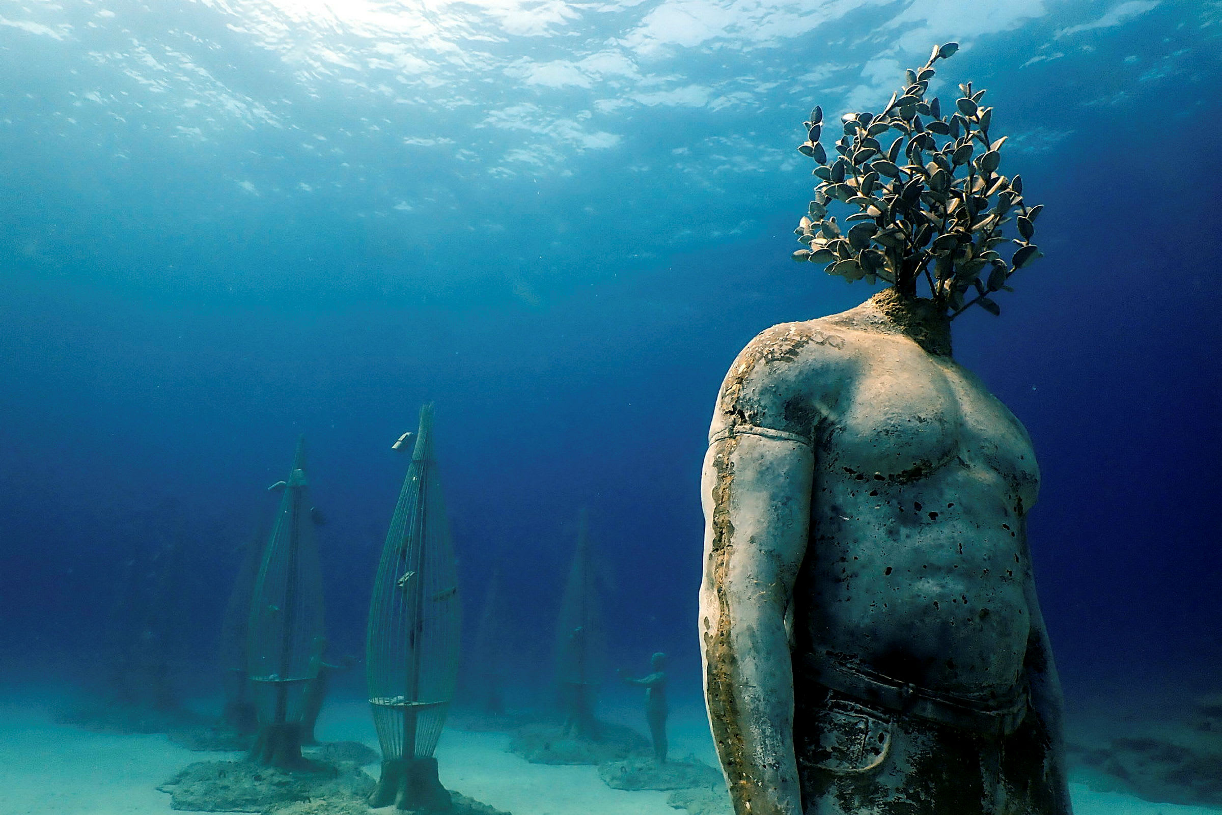 Undervandsbillede med en menneskelig skulptur, hvor hovedet er erstattet af vegetation. Bag statuen ses flere strukturer, og der anes lys ovenfor. (AI-genereret tekst)