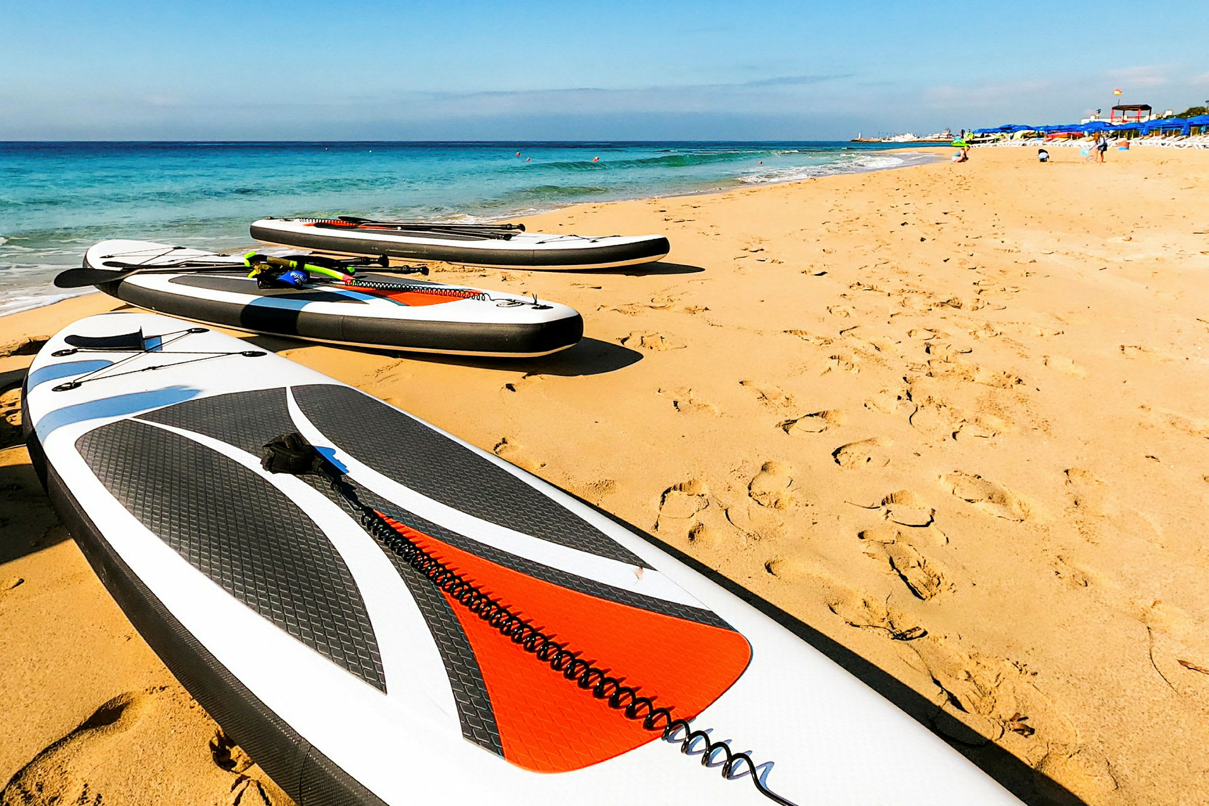 Billede af en sandstrand med flere stand up paddleboards placeret på sanden tæt på vandet. (AI-genereret tekst)