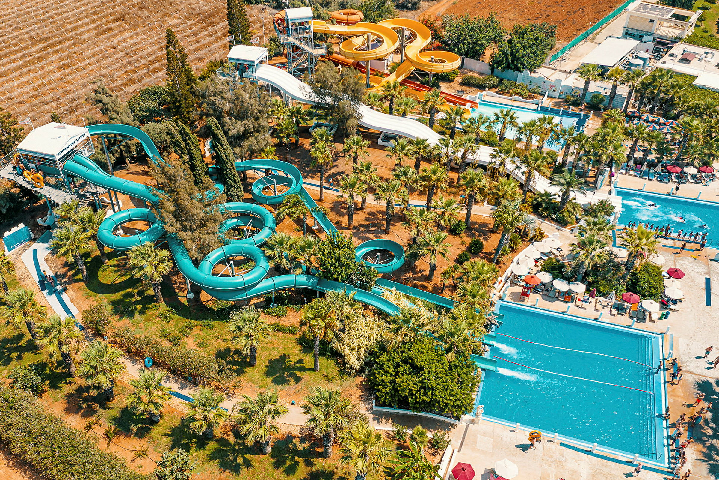 Luftfoto af en vandpark med pools, vandrutsjebaner og omkringliggende træer og vegetation. (AI-genereret tekst)