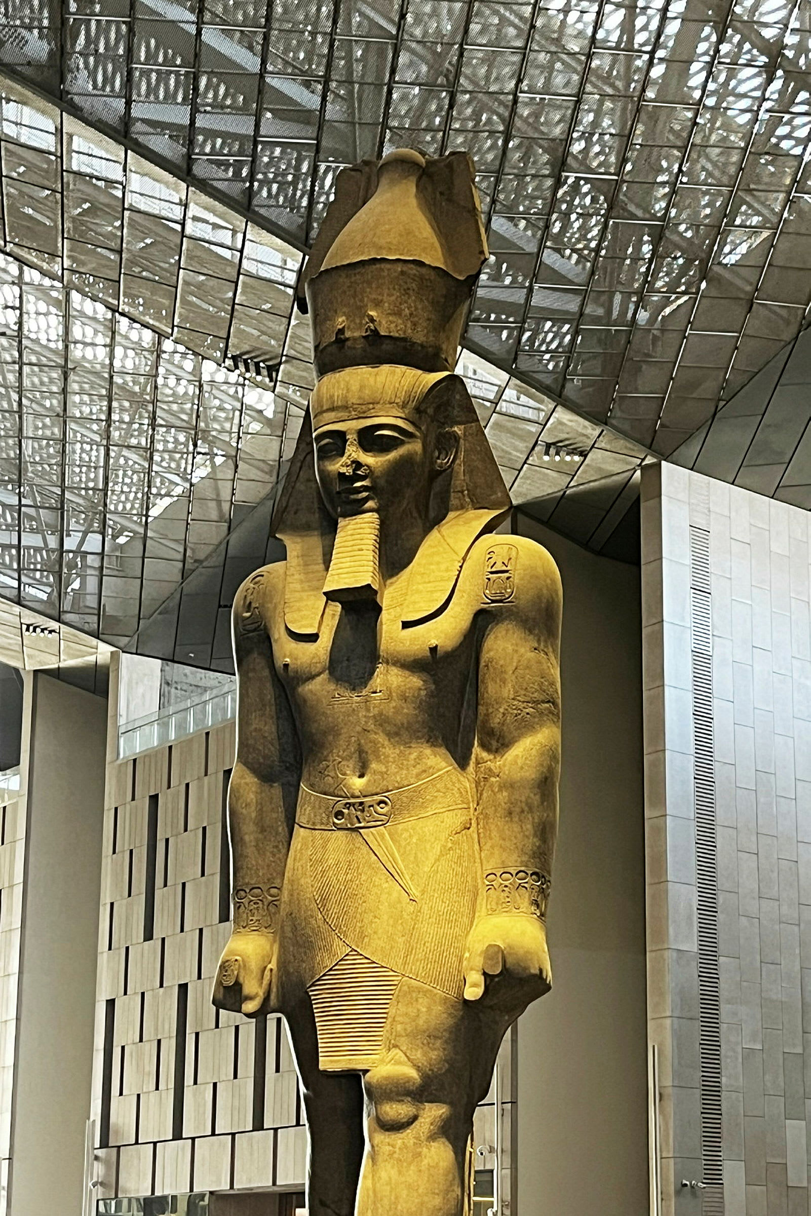 En stor antik skulptur af en stående mandefigur med detaljeret påklædning og hovedbeklædning, placeret i en moderne bygning. (AI-genereret tekst)