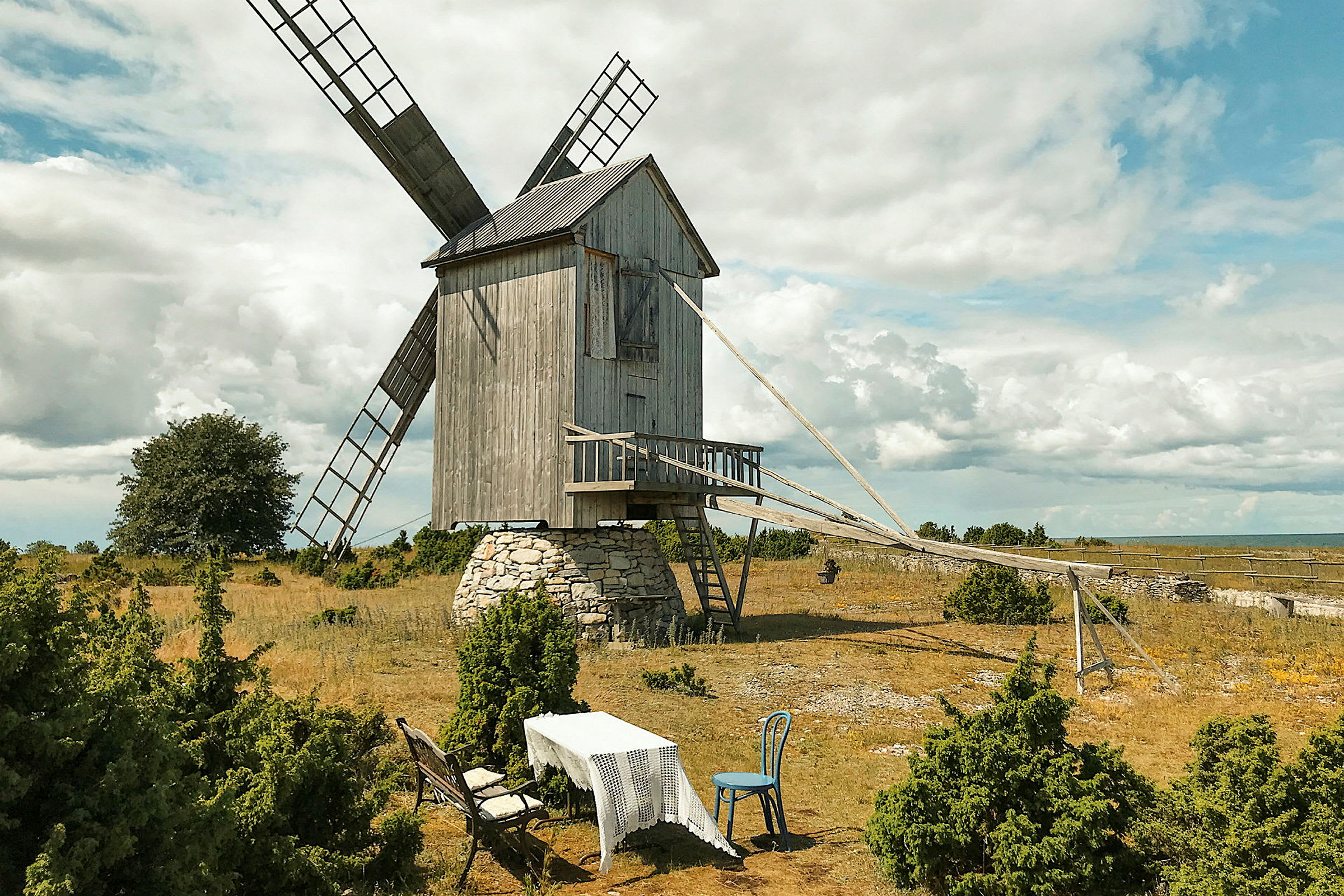 En gammel træmølle på en stenfod i et åbent landskab med græs og buske med et bord og stole foran. (AI-genereret tekst)