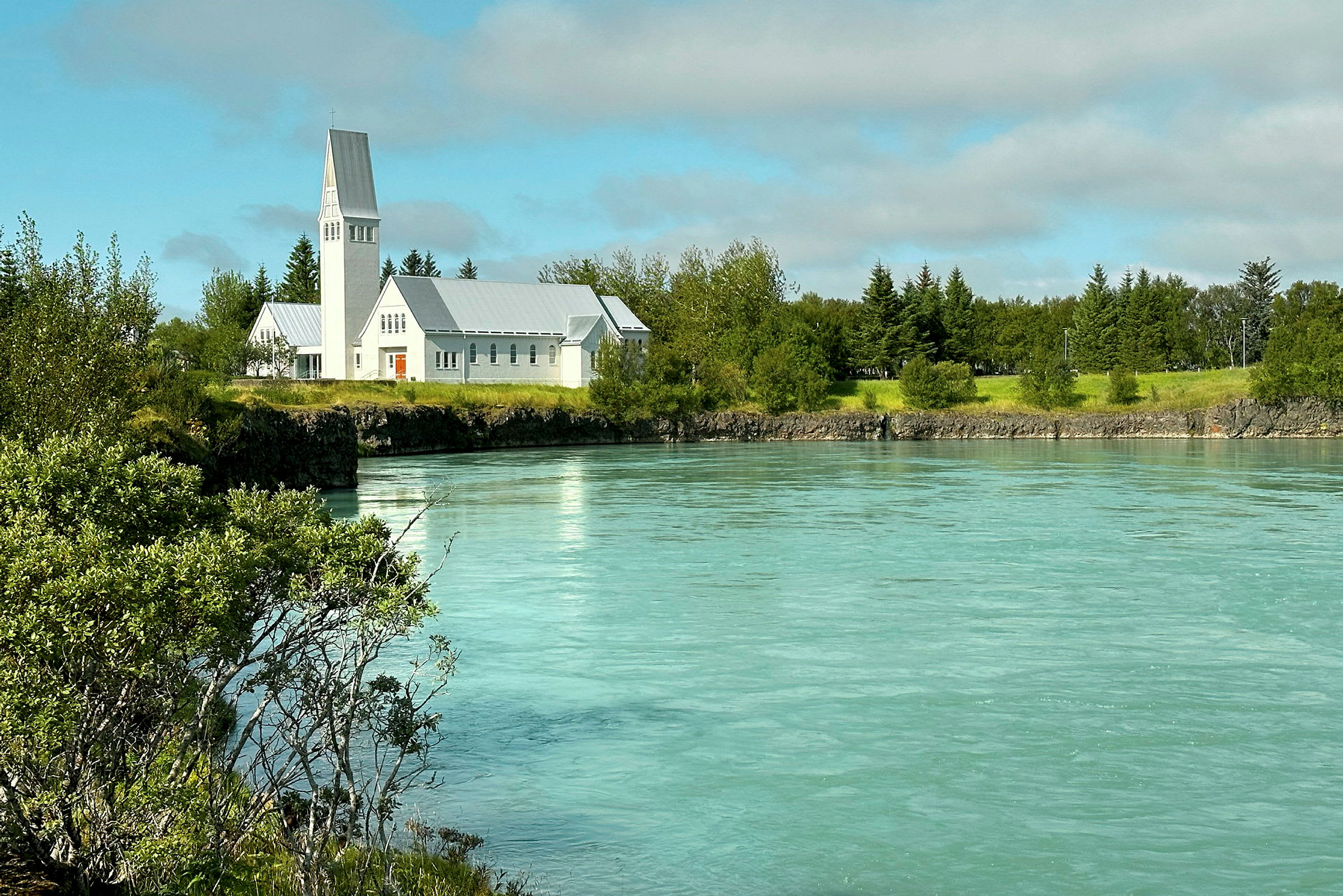 En hvid kirke ligger ved en turkis flod omgivet af grønt og træer. (AI-genereret tekst)