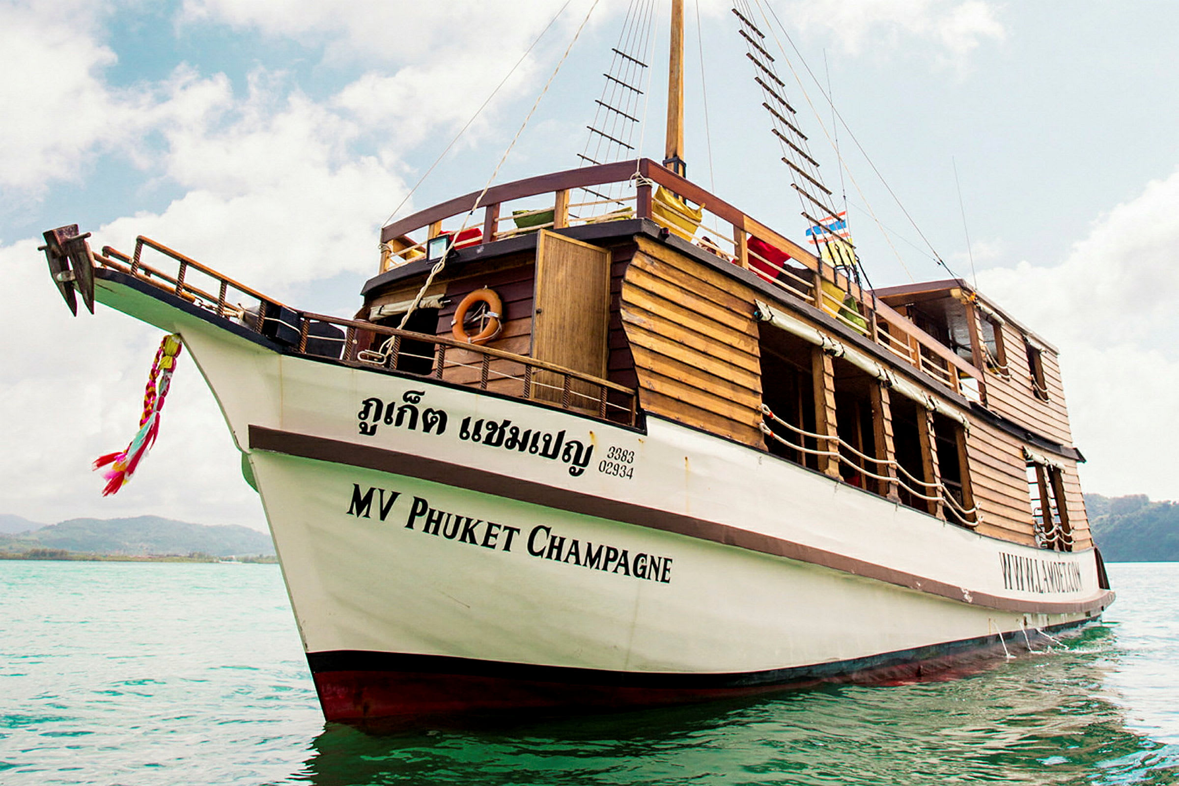 Et træskib med teksten 'MV Phuket Champagne' på skroget er forankret i klart vand. (AI-genereret tekst)