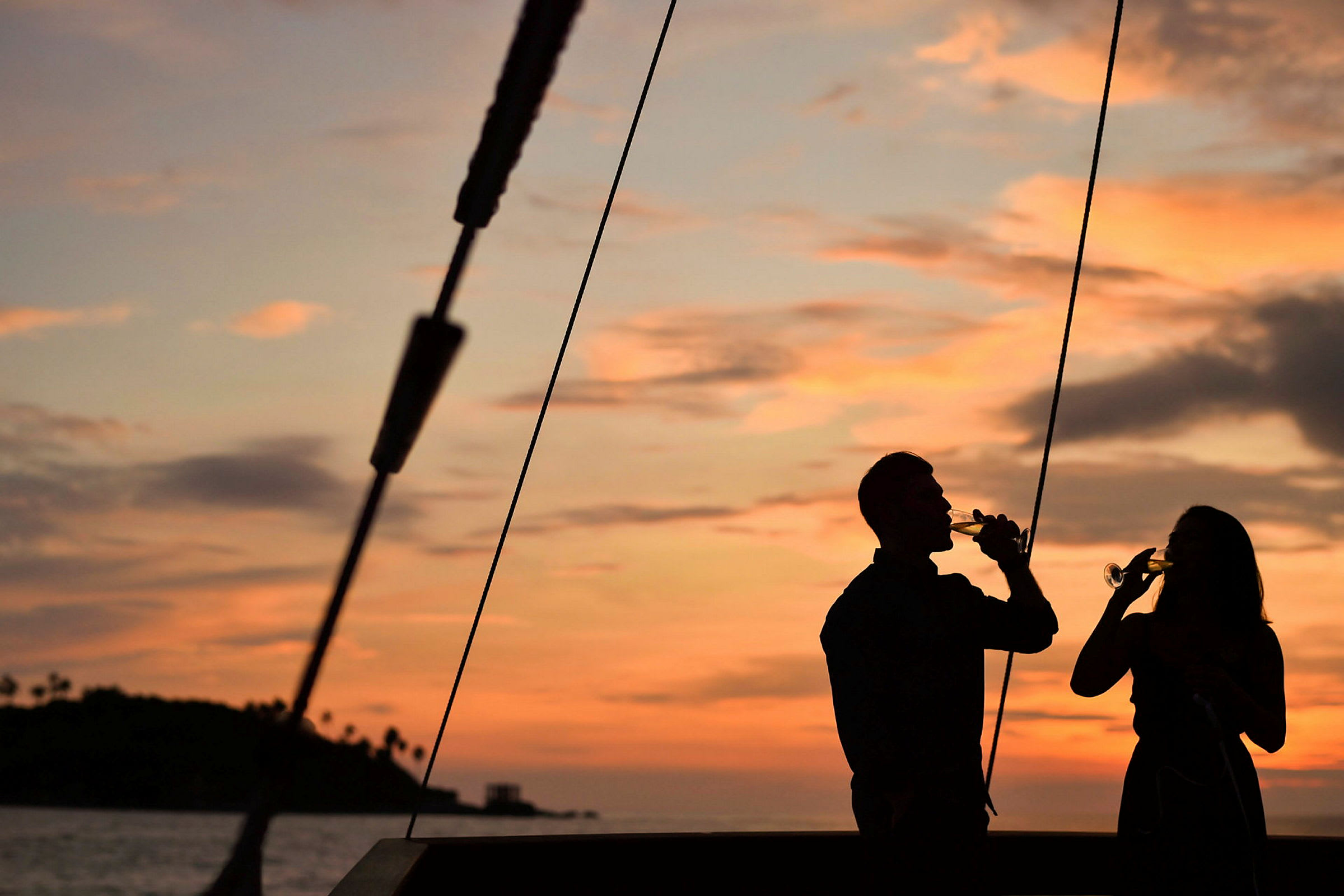 Silhuetter af to personer, der holder glas ved solnedgang med havet og en lille ø i baggrunden. (AI-genereret tekst)