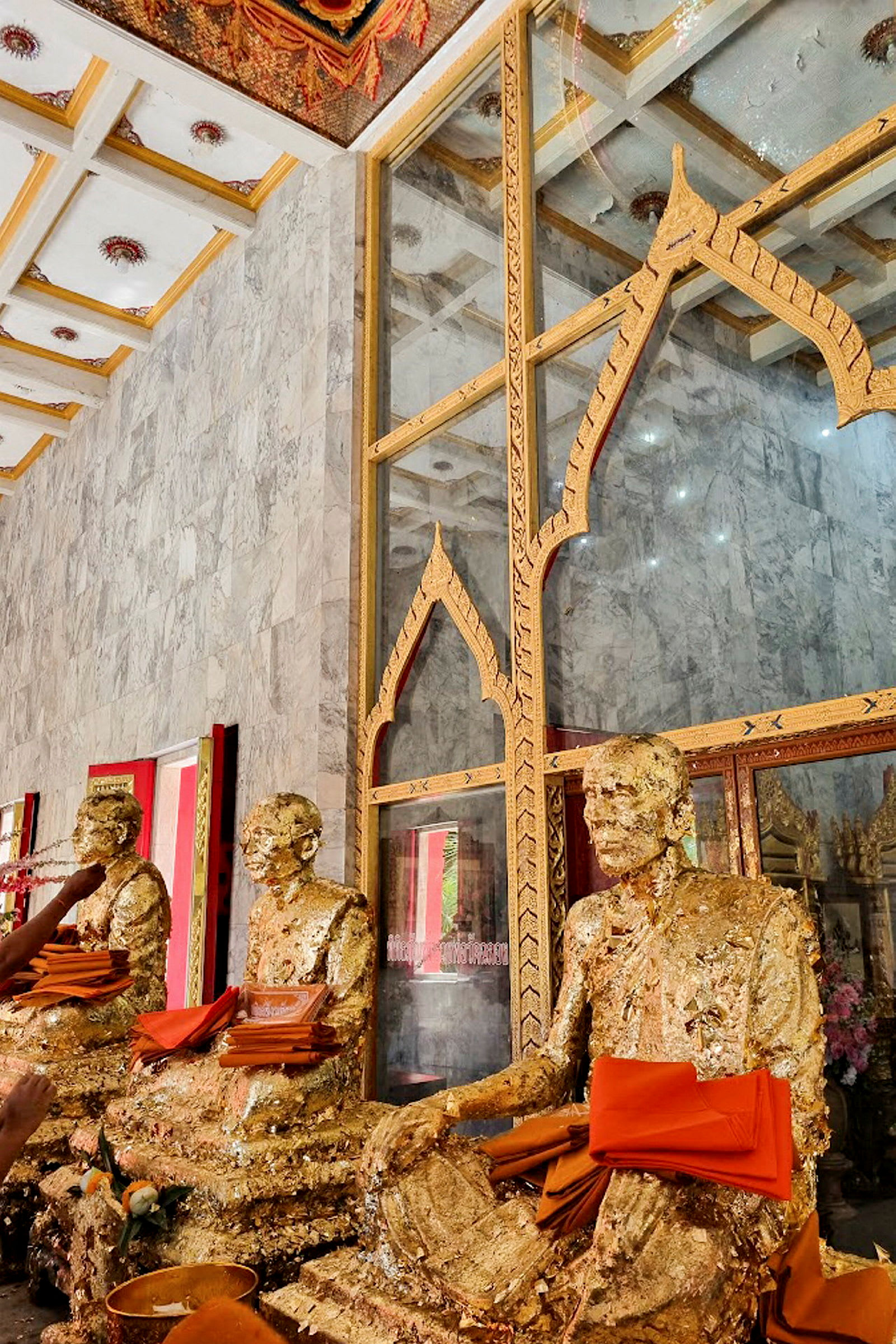 Billede af guldfarvede buddhistiske statuer dekoreret med orange tekstiler foran en glasvæg med udsmykninger. (AI-genereret tekst)