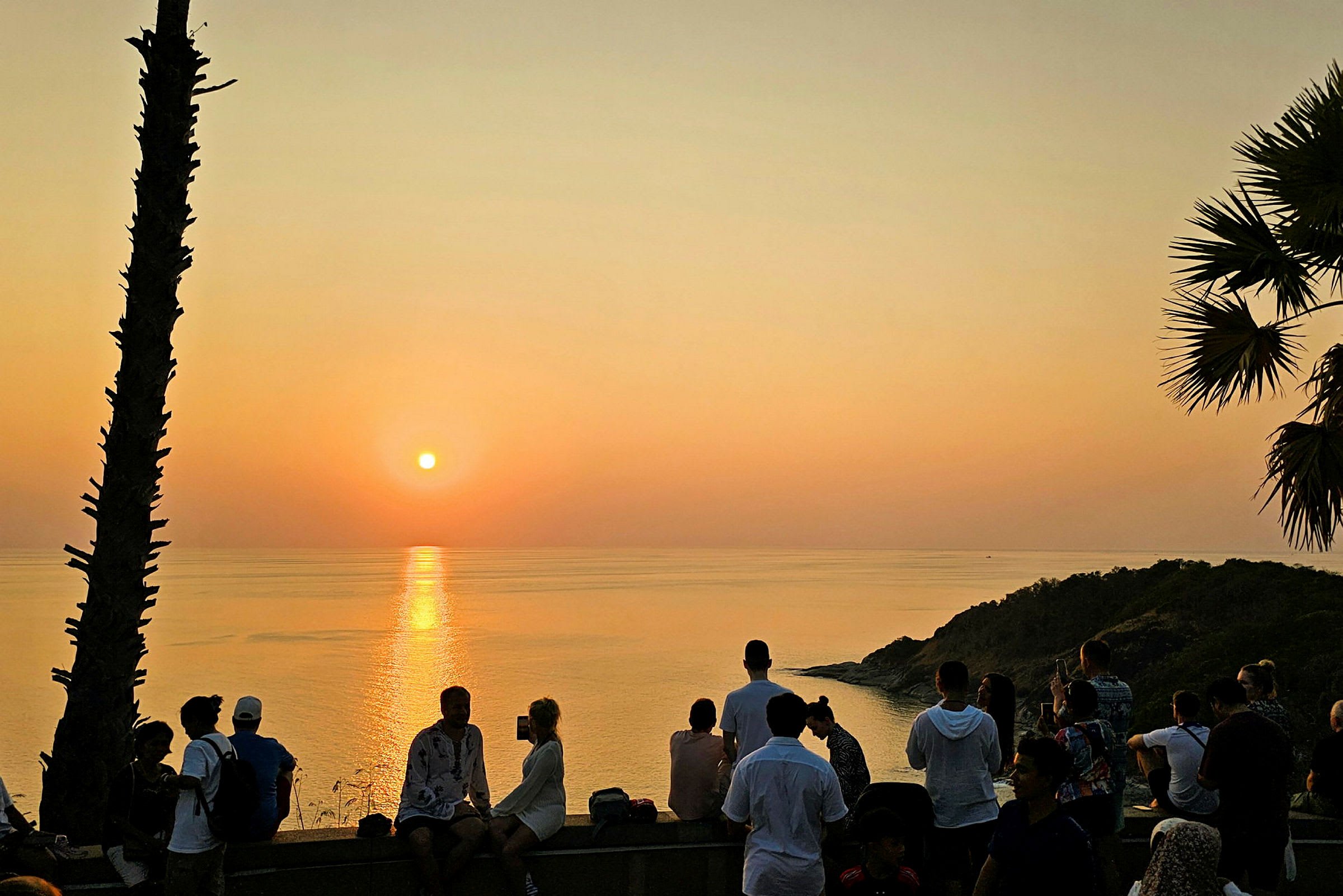 Solnedgang over havet med folk samlet på et udsigtspunkt. (AI-genereret tekst)